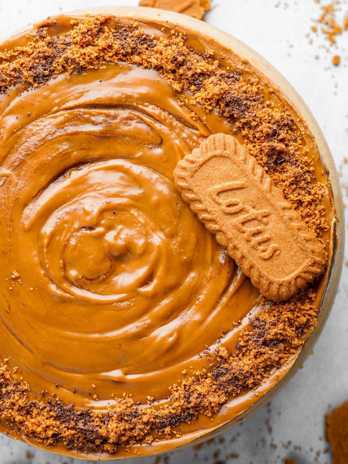 Overhead view of a cake topped with smooth caramel-colored frosting, crumbled biscuits, and a whole Lotus Biscoff biscuit for decoration.