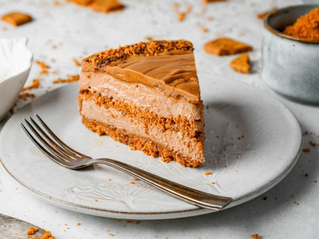 A slice of layered caramel and cookie cheesecake sits on a white plate with a fork, surrounded by cookie crumbs.