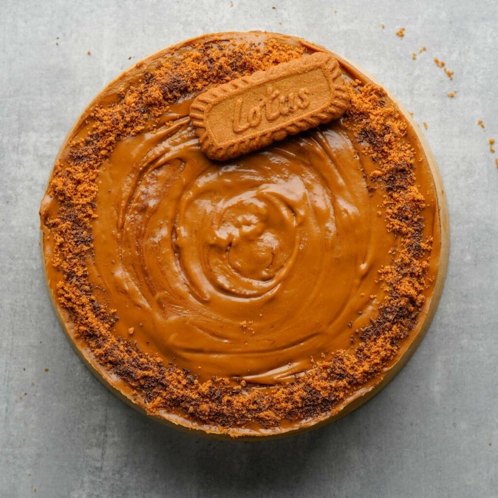 A round cake topped with smooth caramel-colored spread, crushed cookie crumbs around the edge, and a whole Lotus Biscoff cookie placed on top.