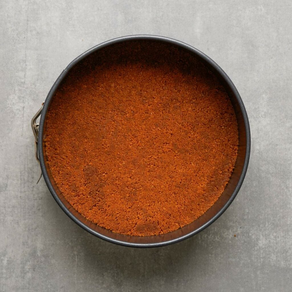 A round baking pan filled with an even layer of crushed graham cracker crust mixture, placed on a gray surface.