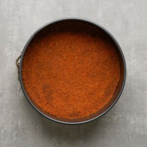 A round baking pan filled with an even layer of crushed graham cracker crust mixture, placed on a gray surface.