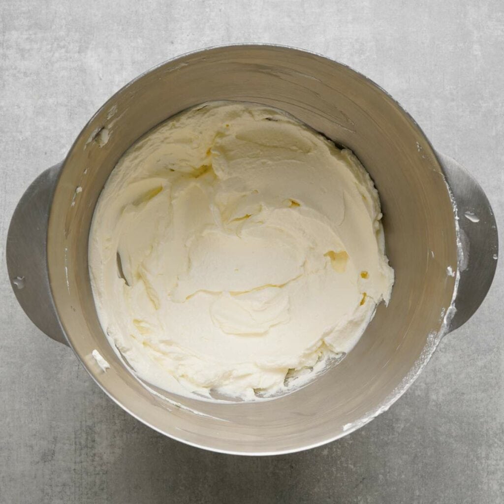 A metal mixing bowl filled with smooth, whipped cream cheese frosting on a gray surface.
