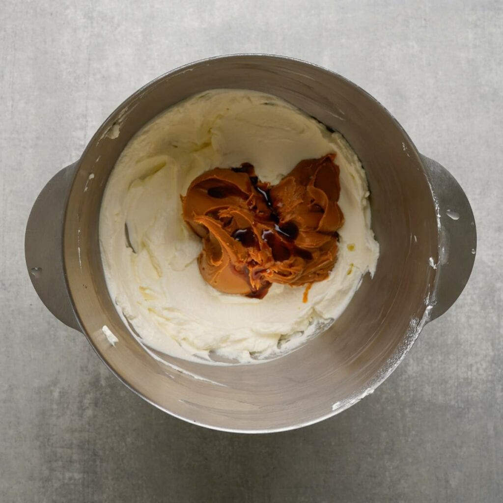 A metal mixing bowl containing whipped cream cheese, Biscoff spread, and vanilla extract on a gray surface.