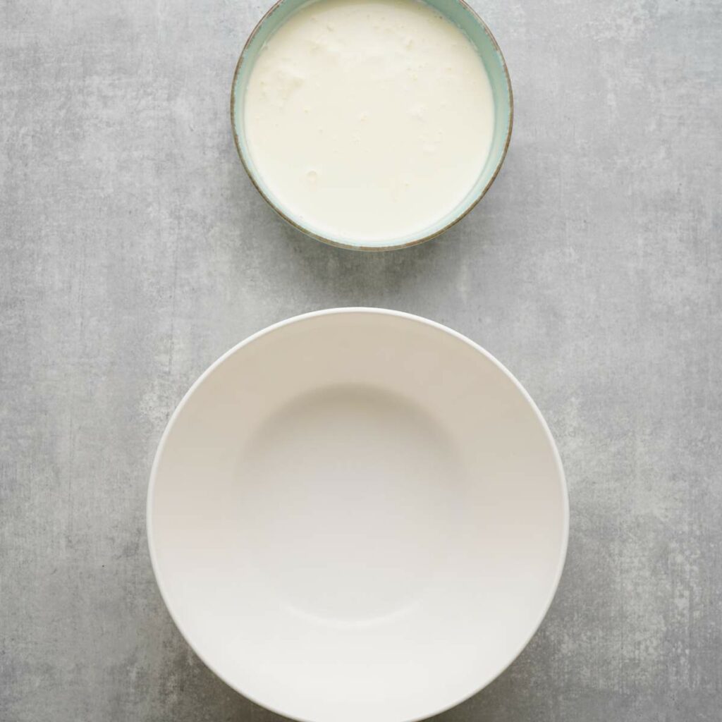 A small bowl filled with a creamy white liquid is placed above an empty, larger white bowl on a gray surface.