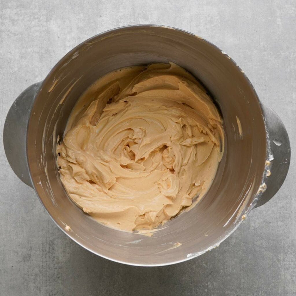 A metal mixing bowl filled with light brown creamy batter or frosting, set on a gray countertop.