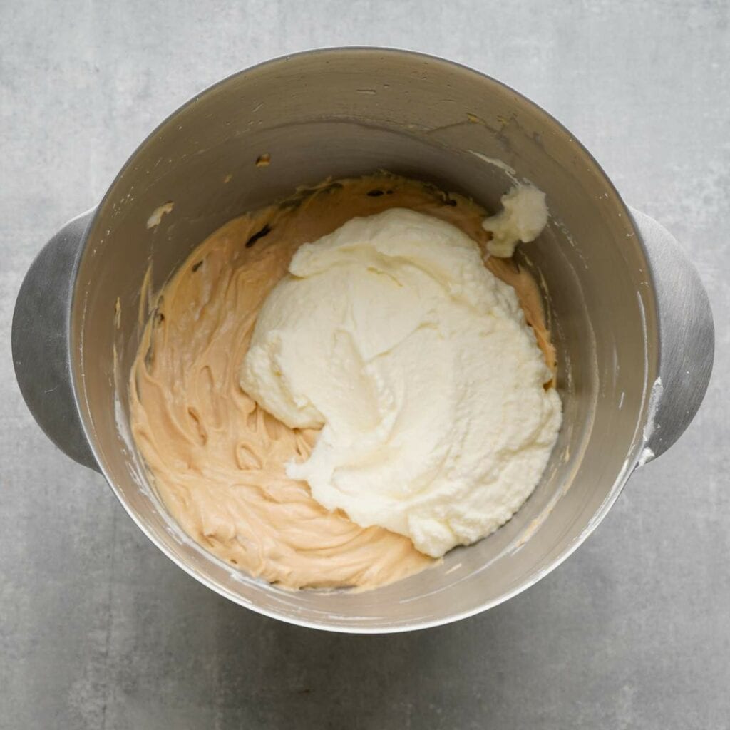 A metal mixing bowl contains two different mixtures: a light tan batter and a white whipped mixture, partially combined.