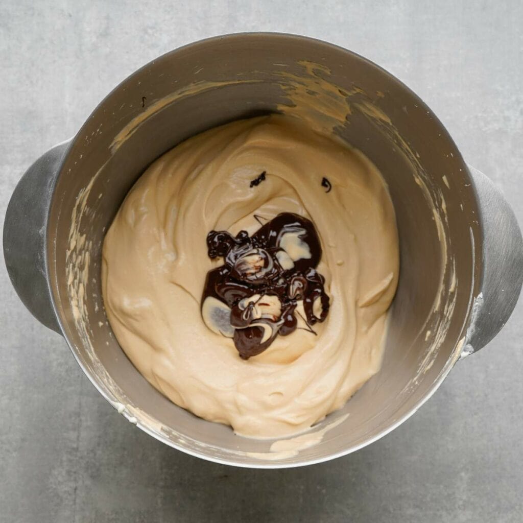 A mixing bowl with creamy batter and melted chocolate partially blended on top, set on a gray surface.