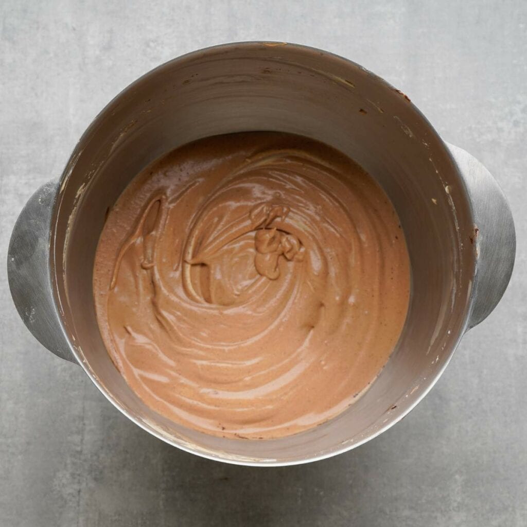 A metal mixing bowl containing smooth, swirled chocolate cake batter on a gray surface.