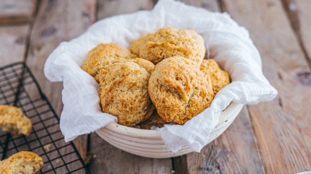 A white bowl lined with a cloth holds several freshly baked buterrmilk biscuits.