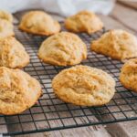 Golden brown drop biscuits cooling on a black wire rack placed on a wooden surface, with a white cloth in the background.