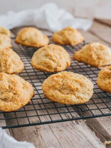 Golden brown drop biscuits cooling on a black wire rack placed on a wooden surface, with a white cloth in the background.