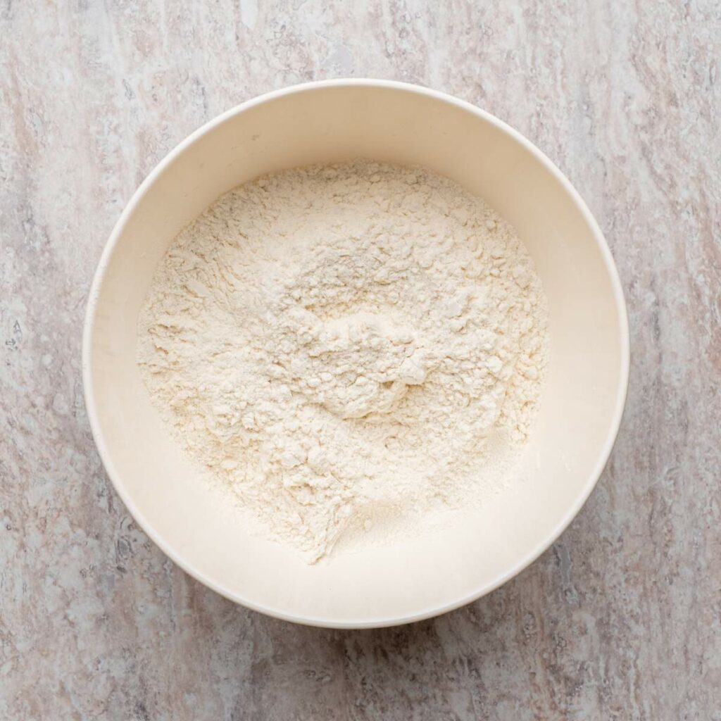A white mixing bowl filled with dry flour sits on a light-colored marble countertop.