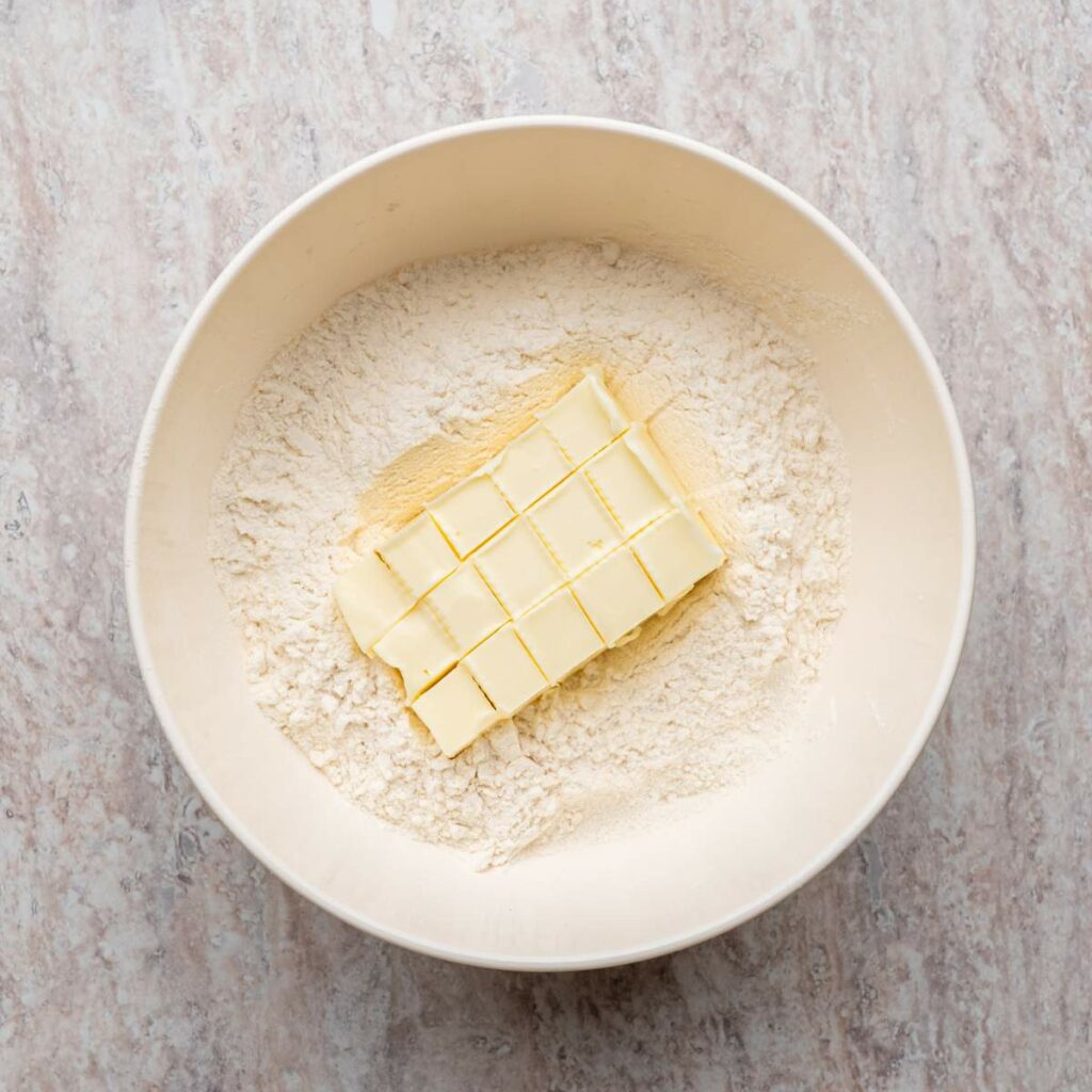 Cubes of butter placed on top of flour in a large mixing bowl, viewed from above on a light countertop.
