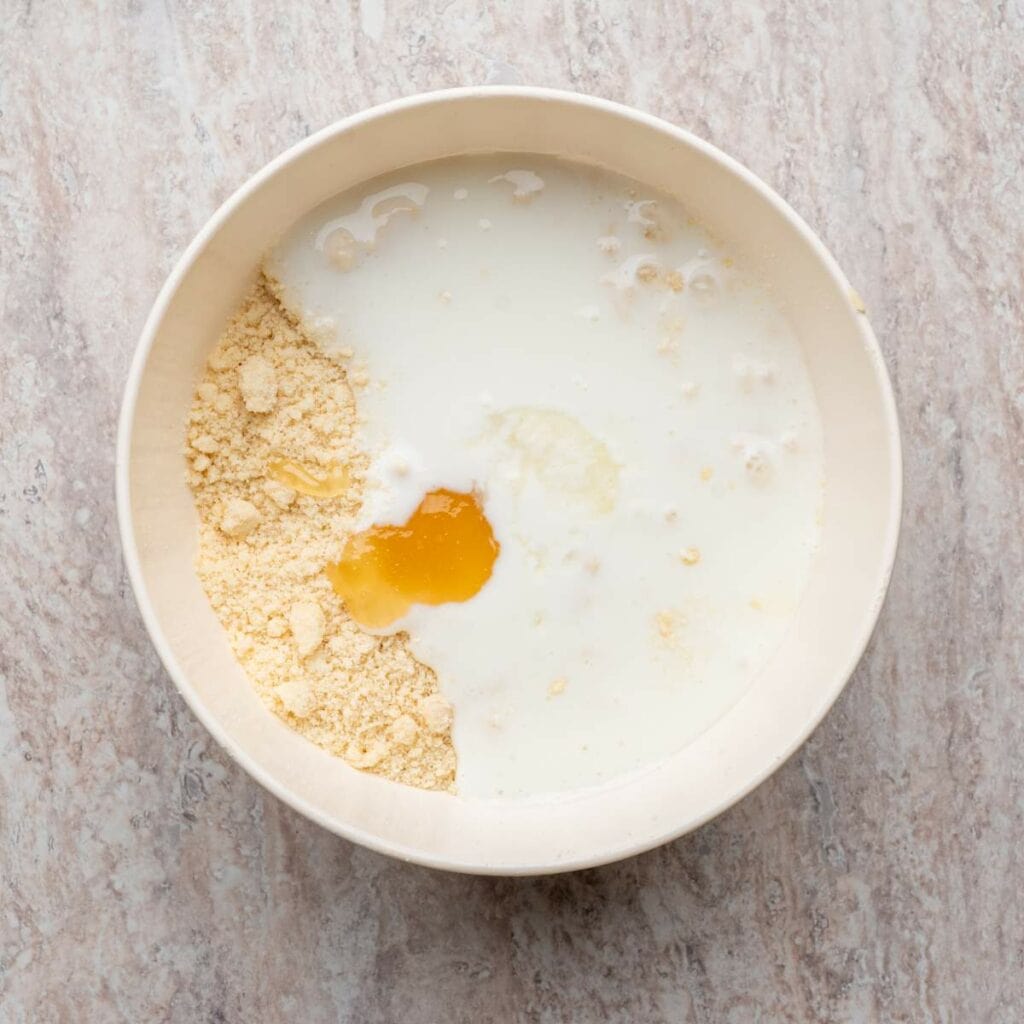 A bowl containing flour, milk, and a cracked egg on a light-colored surface.
