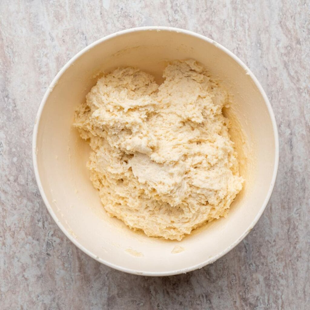 A white bowl filled with raw, mixed dough sits on a light-colored, textured surface.