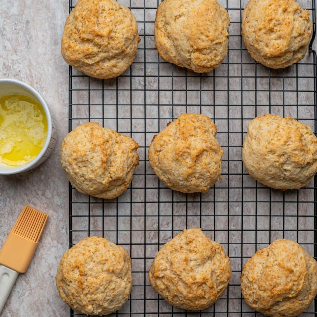 Nine golden brown biscuits cooling on a black wire rack, with a bowl of melted butter and a pastry brush placed to the left.