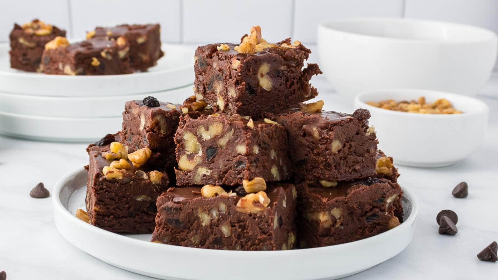 A plate stacked with squares of chocolate fudge, with more pieces and bowls of nuts in the background.