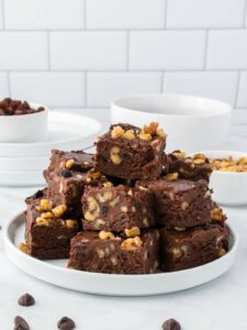 A plate stacked with chocolate fudge containing walnuts, set on a white countertop with bowls and plates in the background.