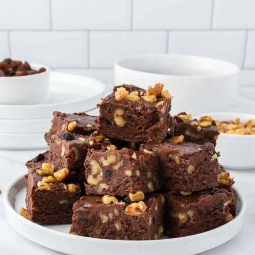 A plate stacked with chocolate fudge containing walnuts, set on a white countertop with bowls and plates in the background.