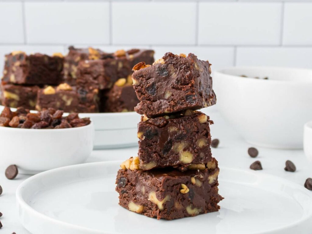 Three pieces of chocolate fudge with nuts are stacked on a white plate, with bowls of raisins and chocolate chips in the background.