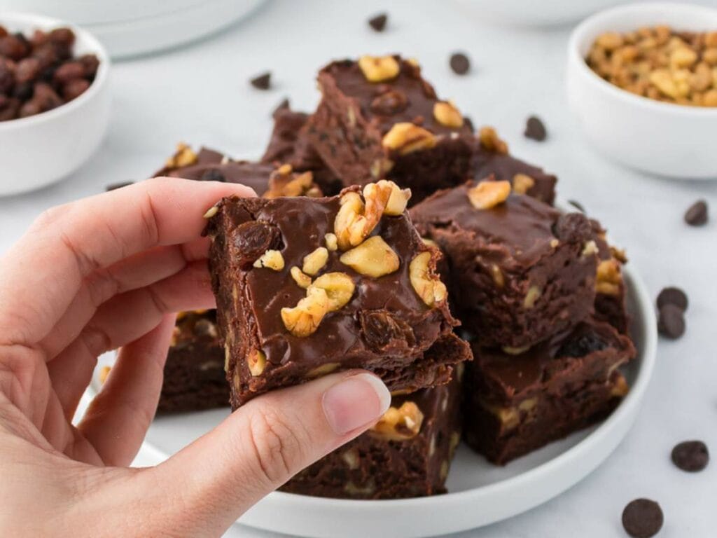 A hand holding a square piece of chocolate fudge with walnuts, with more pieces of fudge stacked on a white plate.