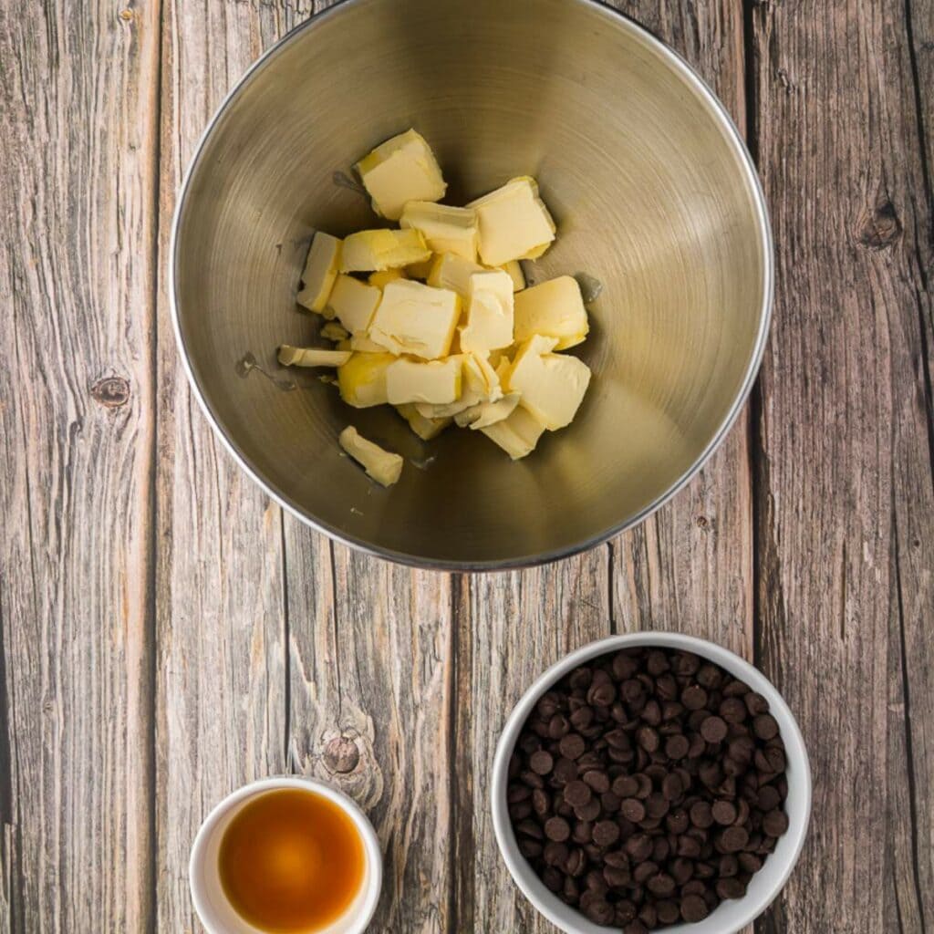 A metal bowl with sliced butter, a white bowl of chocolate chips, and a small bowl of vanilla extract on a wooden surface.
