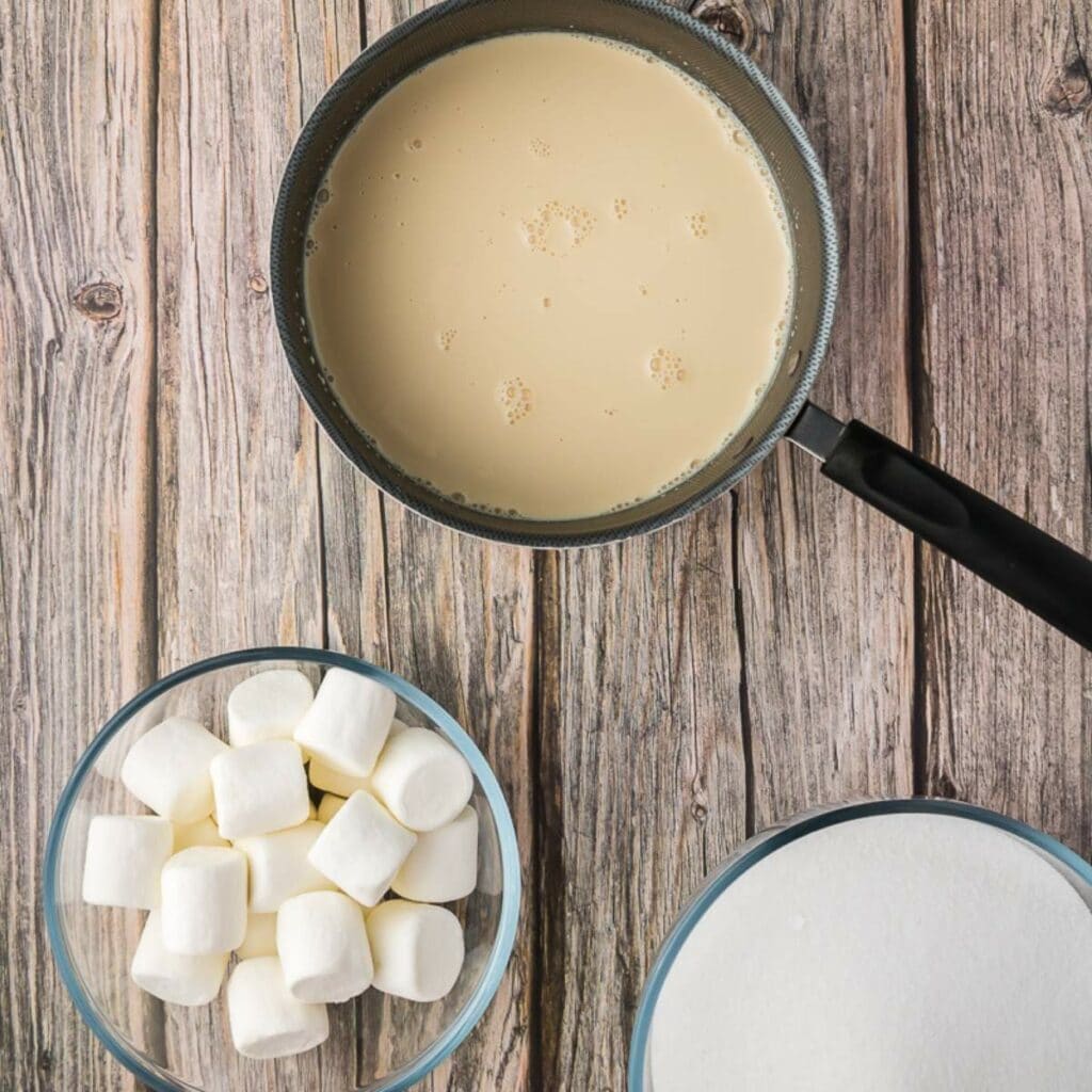 A saucepan with a creamy liquid, a bowl of large marshmallows, and a bowl of white granulated sugar on a wooden surface.