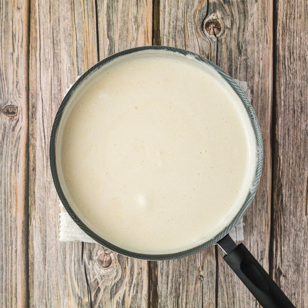 A saucepan filled with a light-colored creamy sauce sits on a rustic wooden surface.