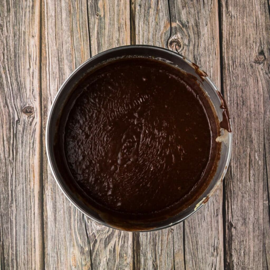 A round baking pan filled with chocolate batter sits on a rustic wooden surface.