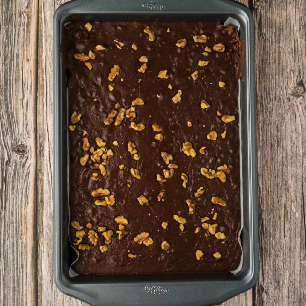 A baking pan filled with unbaked fudge batter topped with chopped walnuts, placed on a wooden surface.