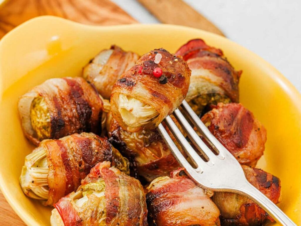 A fork holds a bacon-wrapped Brussels sprout above a yellow dish filled with more bacon-wrapped Brussels sprouts.