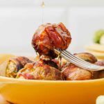 A fork holds a bacon-wrapped Brussels sprout above a yellow dish, with honey being drizzled on top.