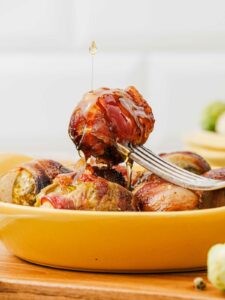A fork holds a bacon-wrapped Brussels sprout above a yellow dish, with honey being drizzled on top.