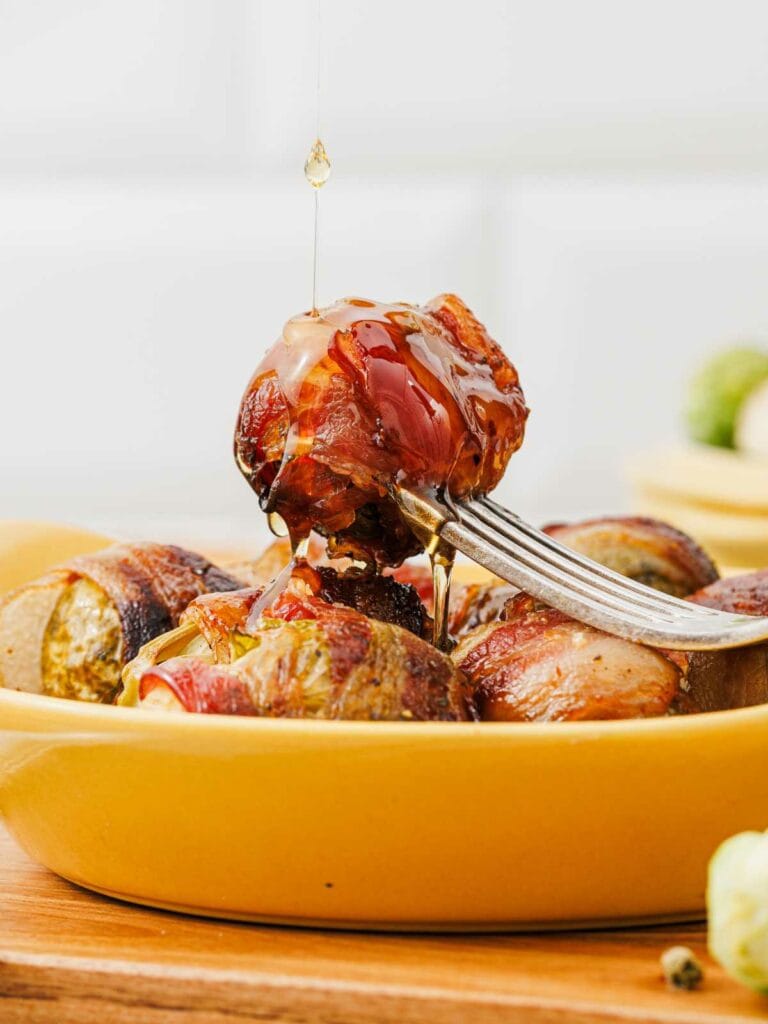A fork holds a bacon-wrapped Brussels sprout above a yellow dish, with honey being drizzled on top.