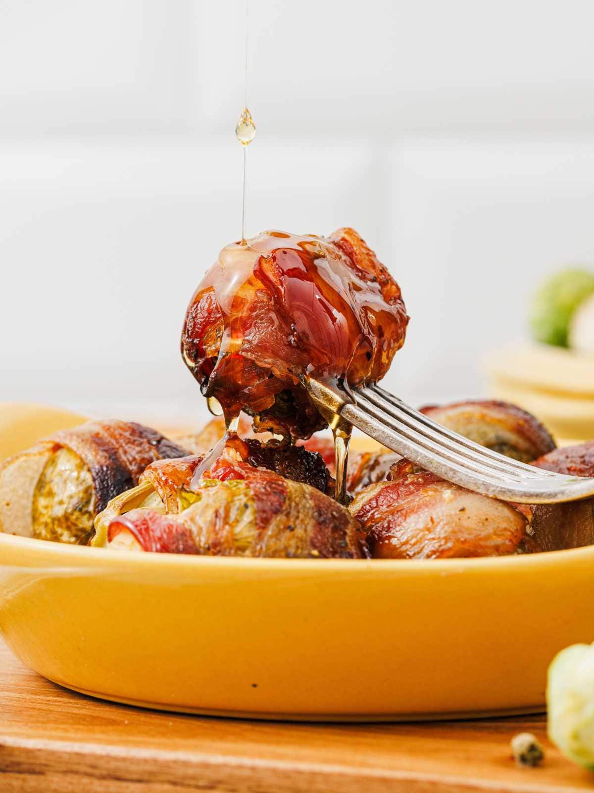 A fork holds a bacon-wrapped Brussels sprout above a yellow dish, with honey being drizzled on top.