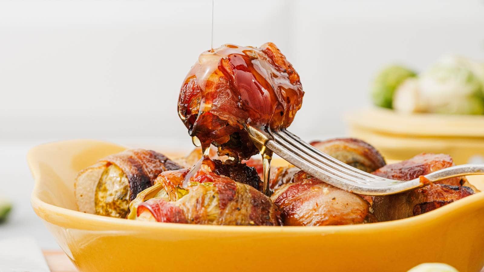 A fork holds a bacon-wrapped Brussels sprout being drizzled with honey over a dish filled with more bacon-wrapped Brussels sprouts.
