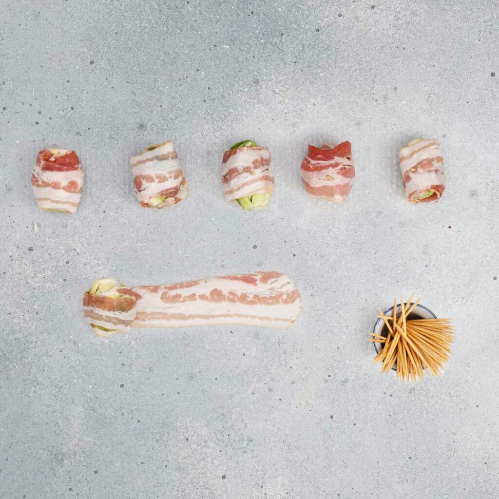 Six pieces of bacon-wrapped food, one unwrapped strip with filling, and a pile of toothpicks on a gray surface.