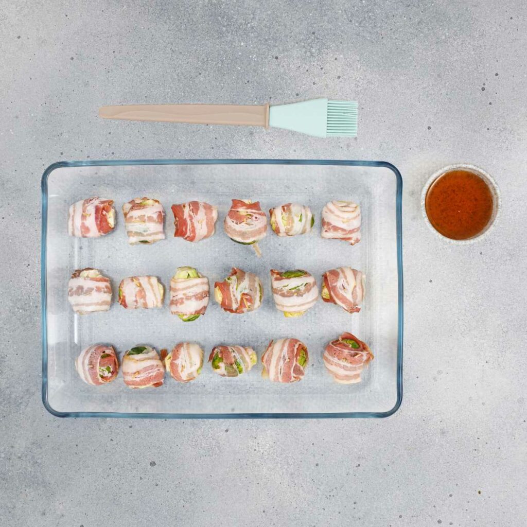 A glass baking dish with rows of bacon-wrapped Brussel sprouts, a silicone basting brush, and a bowl of sauce on a gray countertop.