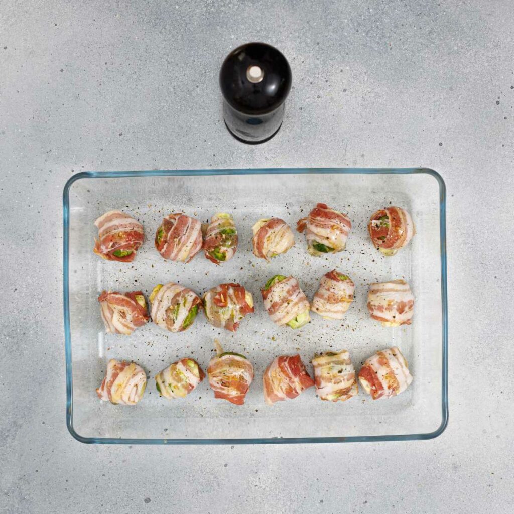 A glass baking dish with bacon-wrapped Brussel sprouts, sprinkled with pepper. A black pepper grinder is positioned above the dish.