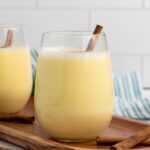 A glass of yellow eggnog with a cinnamon stick, placed on a wooden tray with additional cinnamon sticks nearby.