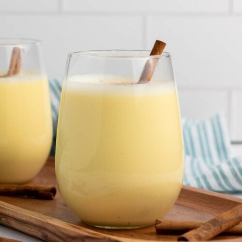 A glass of yellow eggnog with a cinnamon stick, placed on a wooden tray with additional cinnamon sticks nearby.