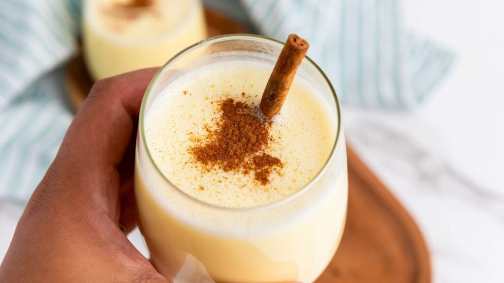 A hand holds a glass of eggnog topped with cinnamon powder and a cinnamon stick.