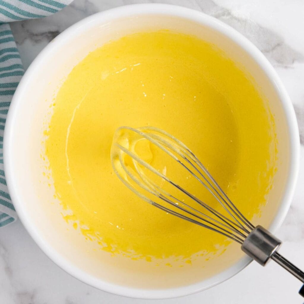 A metal whisk rests in a white bowl containing a smooth, yellow batter.