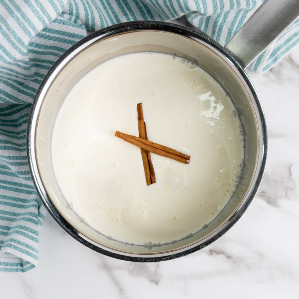 A saucepan filled with milk and two cinnamon sticks on top, placed on a marble surface next to a striped cloth.