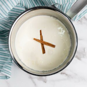 A saucepan filled with milk and two cinnamon sticks on top, placed on a marble surface next to a striped cloth.