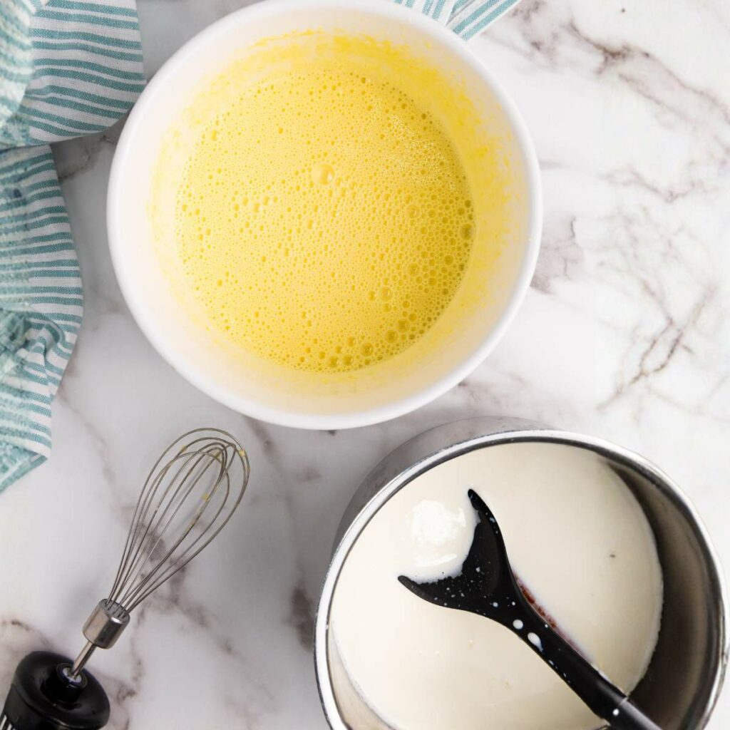 A bowl of whisked yellow mixture beside a pot of white liquid, with a whisk and a black spoon on a marble countertop.