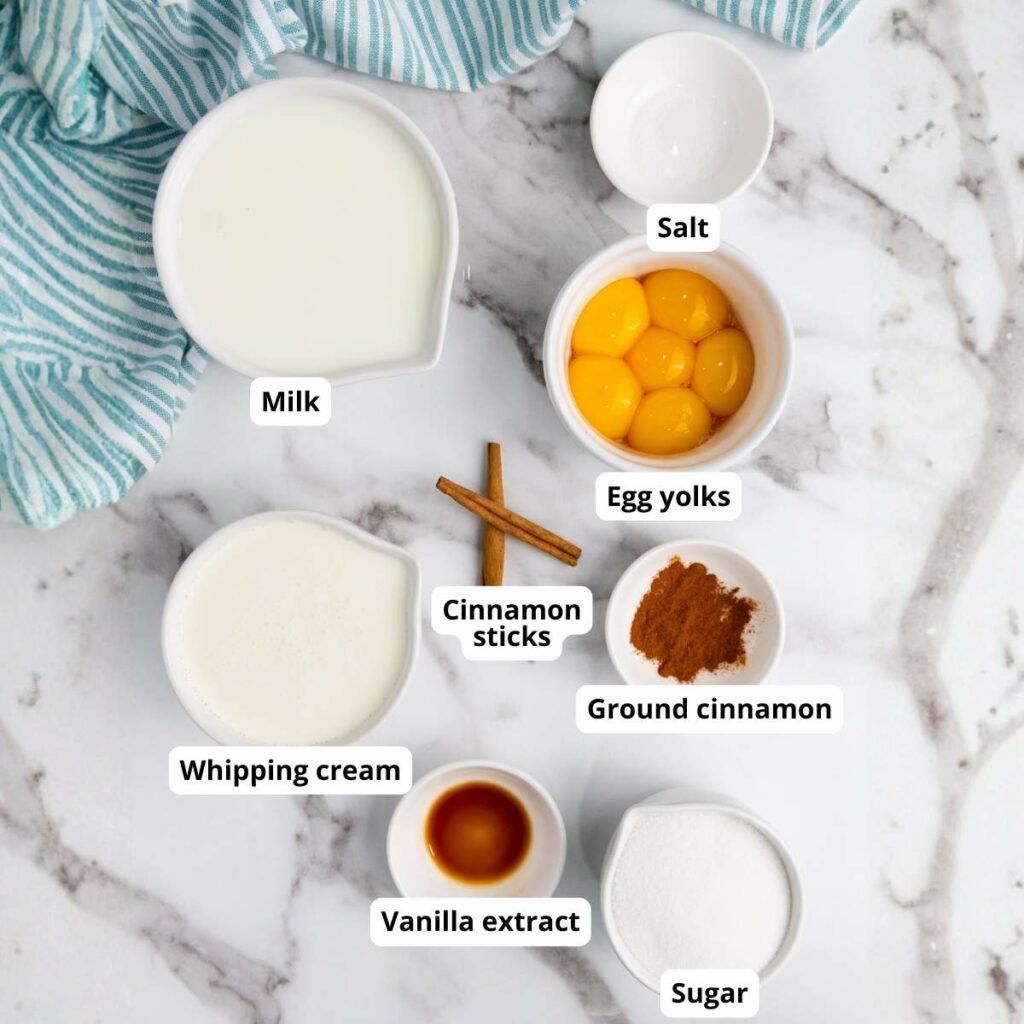A marble surface with bowls containing milk, egg yolks, whipping cream, vanilla extract, salt, cinnamon sticks, ground cinnamon, and sugar.
