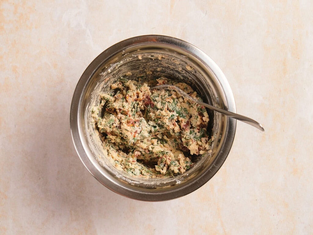 A stainless steel bowl with a spoon containing a mixed filling with herbs and spices on a light-colored surface.
