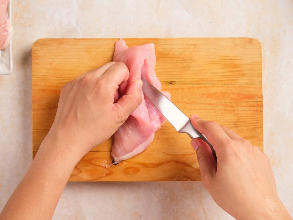 A person uses a knife to cut raw chicken breast on a wooden cutting board.