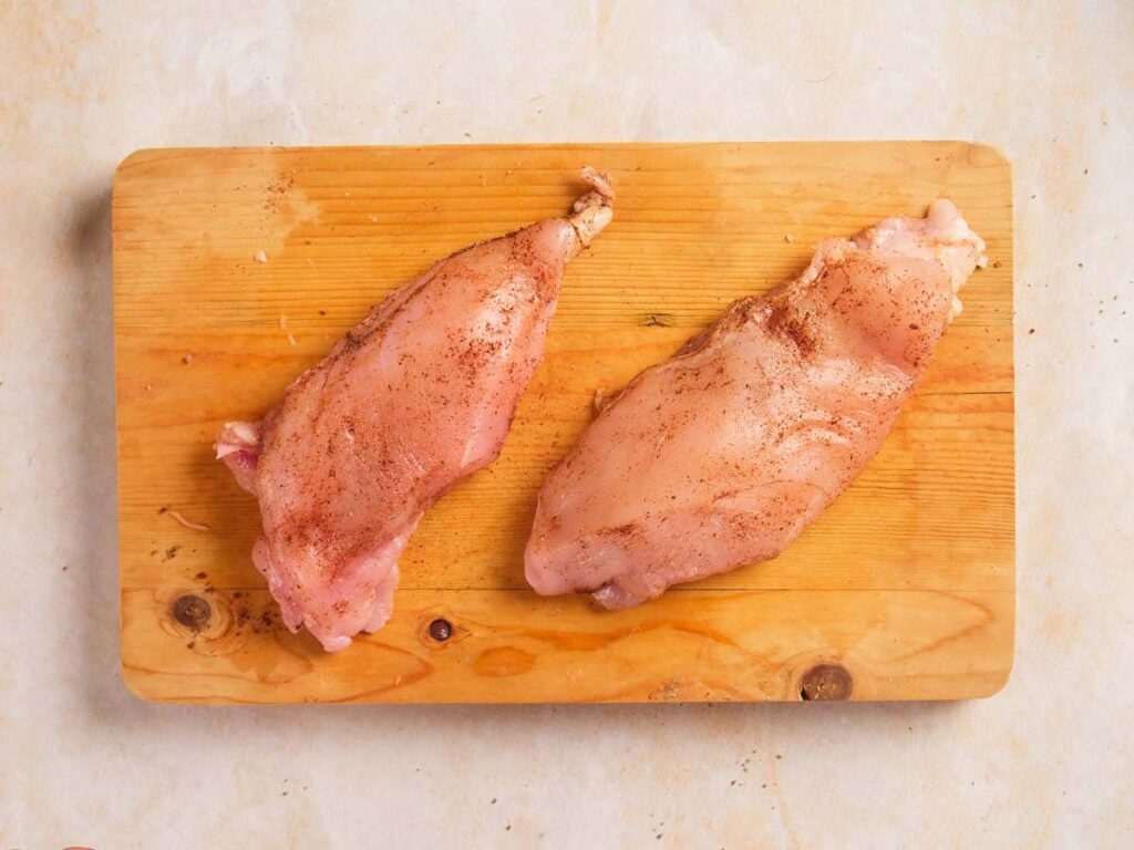 Two raw, seasoned chicken breasts placed on a wooden cutting board.